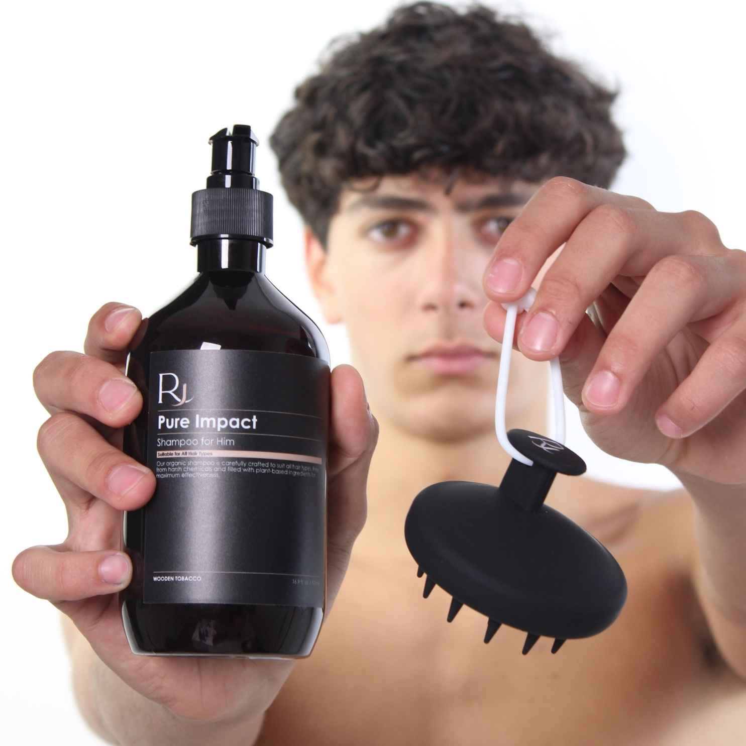 Person holding a bottle of 'Raqiia Pure Impact' shampoo and a black Raqiia scalp massager with a plain background